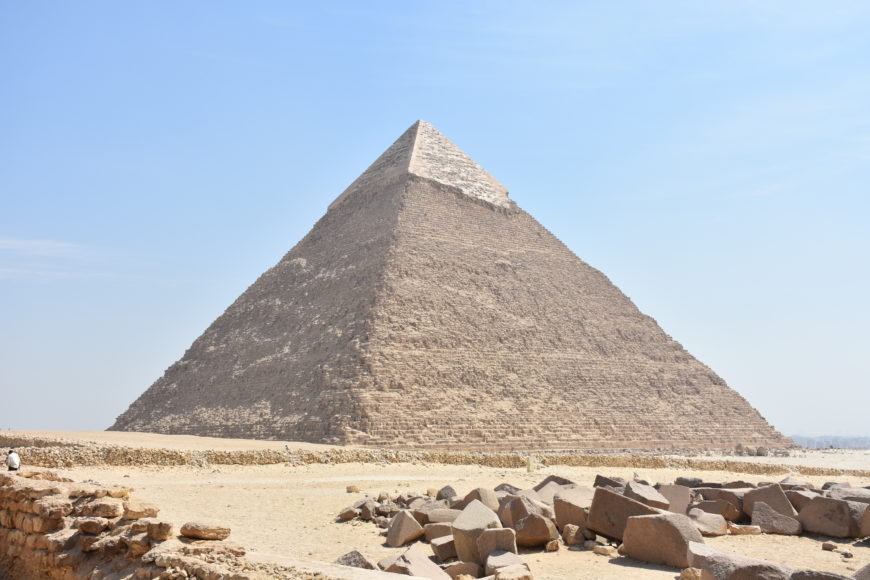 Photograph of the Great Pyramid of Giza showcasing its massive stone structure with a partially preserved smooth limestone casing near the apex. The foreground contains scattered stones and desert terrain under a clear blue sky.