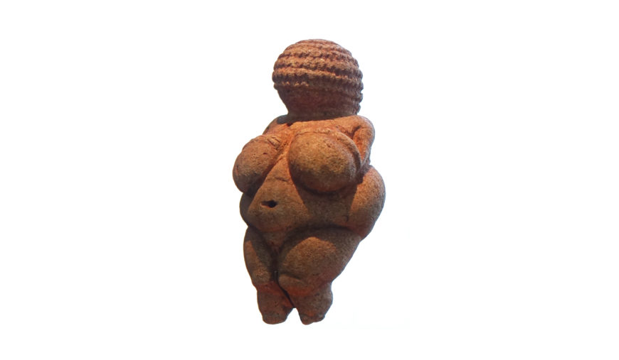 Photograph of a small, ancient figurine known as the Venus of Willendorf, depicting a stylized female form with exaggerated breasts, belly, and hips. The figurine features a textured, braided pattern on the head and lacks facial details, symbolizing fertility.