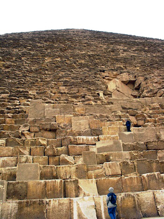Photograph of a large ancient pyramid with two people climbing its massive stone blocks, highlighting scale and structure. The pyramid's weathered stones vary in size and color.