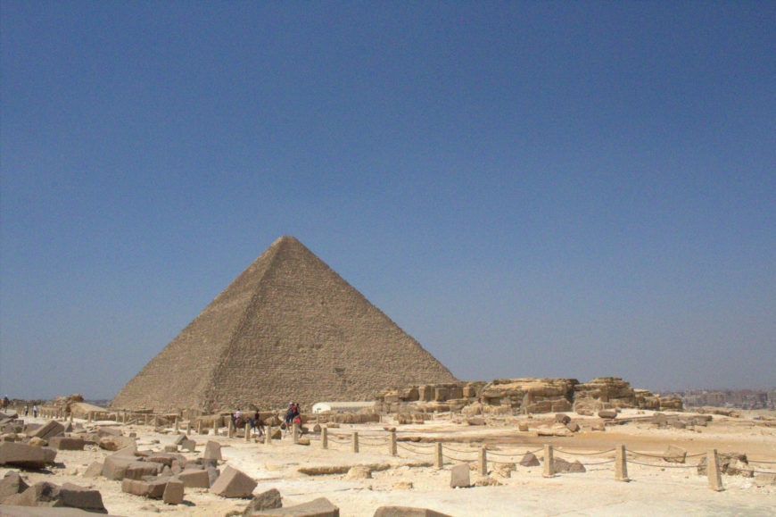 Photograph showing the Great Pyramid of Giza under clear blue sky with scattered ruins and tourists in foreground. The scene highlights ancient Egyptian architecture and archaeological site layout with sandy terrain and stone structures.