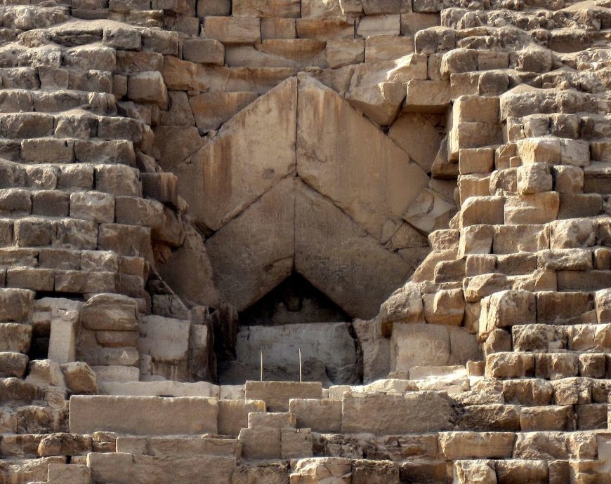Photograph of a close-up section of an ancient Pyramid of Khufu showing a triangular entrance surrounded by weathered, stacked stone blocks. The structure features large, precisely cut stones forming a keystone arch above the entrance, highlighting architectural techniques used in pyramid construction.