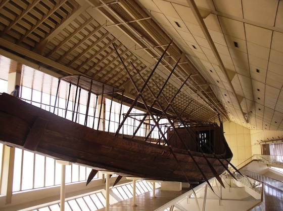 Photograph of an ancient Egyptian wooden boat suspended inside a museum with large windows and a high ceiling. The boat features multiple long wooden poles extending outward, showcasing traditional construction and design elements.