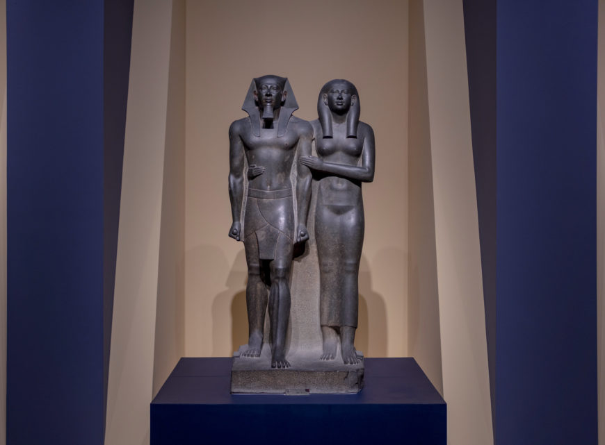 Photograph of a King Menkaure (Mycerinus) and queen statue depicting a standing pharaoh and a woman side by side on a rectangular base. The dark stone sculpture features the pharaoh wearing a traditional headdress and kilt, while the woman wears a long dress with arms crossed, displayed against a neutral background with focused lighting.
