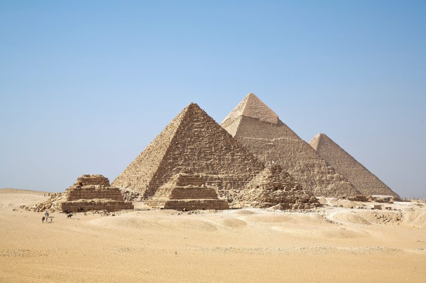 Photograph of Great Pyramids of Giza in desert under clear blue sky, showcasing three large pyramids with smaller ruins in foreground.