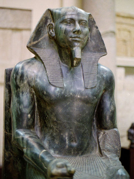 Photograph of an Head and torso (detail), Khafre enthroned statue depicting a seated pharaoh wearing a nemes headdress, carved from dark stone. The statue features detailed musculature and traditional royal attire, highlighting craftsmanship and cultural significance from ancient Egypt.