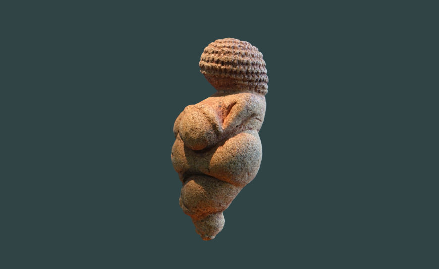 Photograph of the Venus of Willendorf, a small Paleolithic figurine representing a female figure with exaggerated breasts, belly, and hips. The sculpture features detailed braided hair or a woven hat and is believed to symbolize fertility.