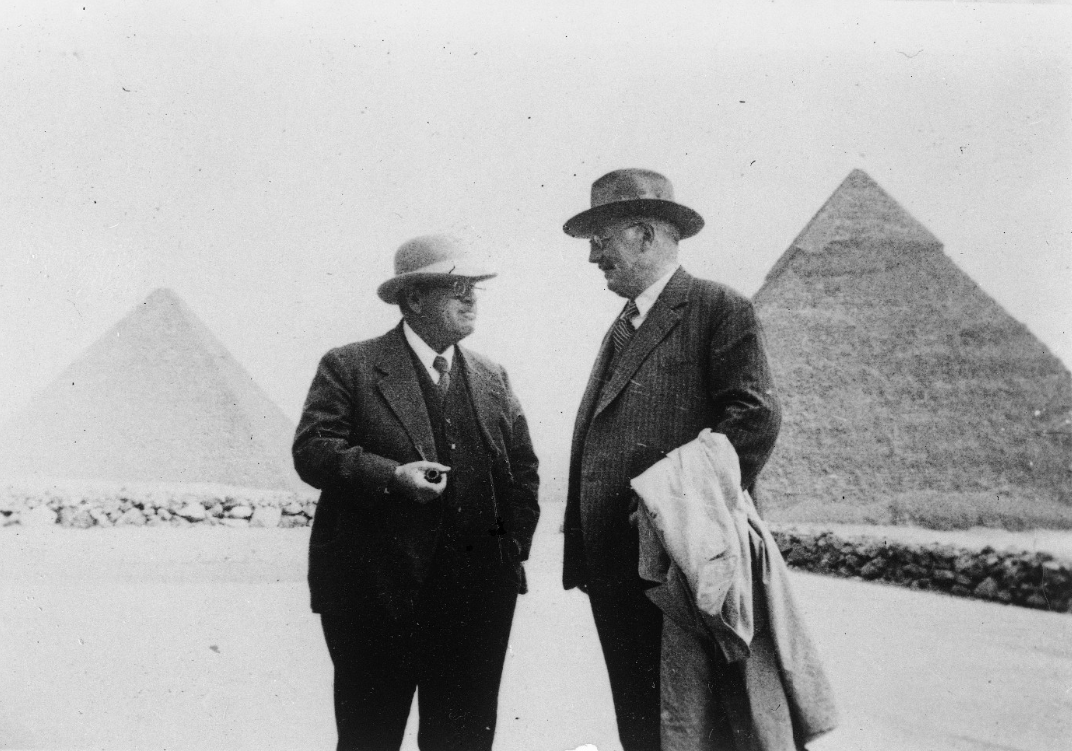 Black and white photograph showing two men in suits and hats standing in front of the Great Pyramids of Giza. The men appear to be engaged in conversation, with one holding a pipe and the other carrying a coat.