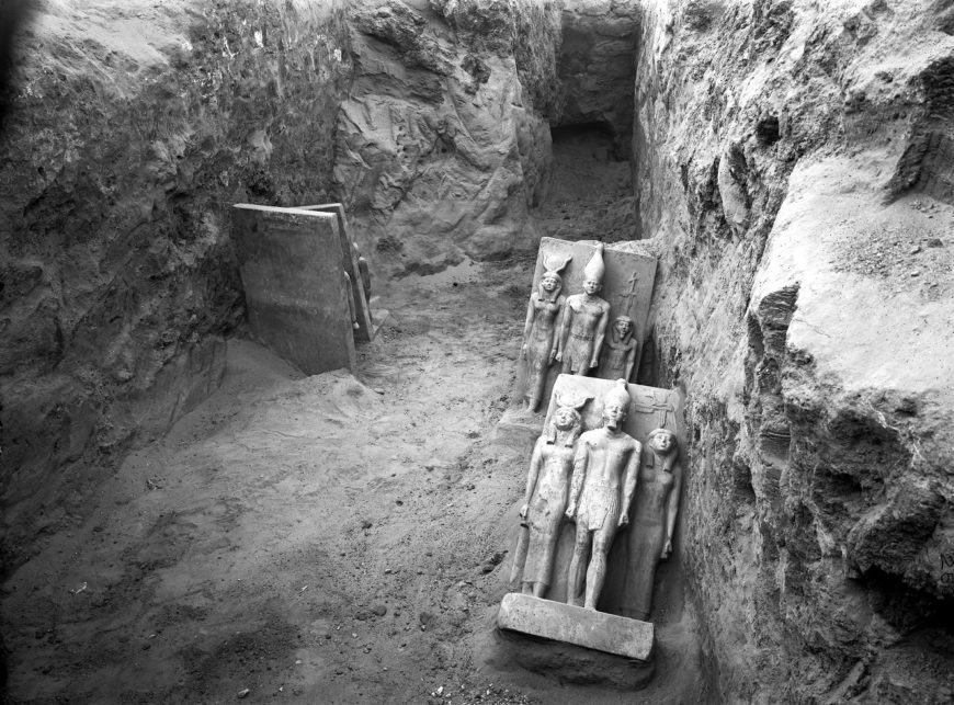Black and white photograph of an archaeological excavation site showing a rectangular pit with four ancient Egyptian statues lying on their backs against the pit walls. The statues depict standing figures wearing traditional headdresses, arranged in two groups, with a partially buried stone slab visible on the left side of the pit.