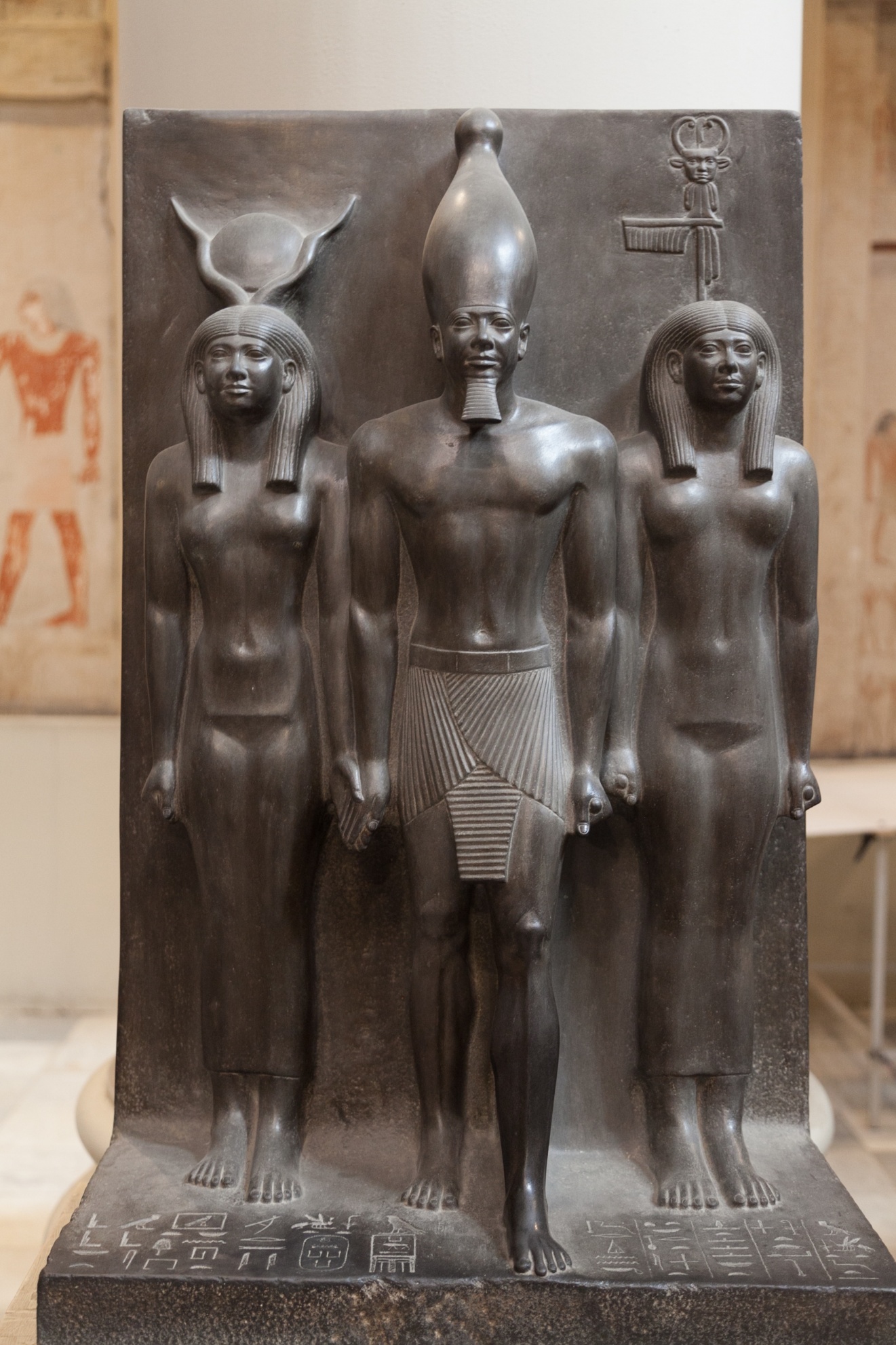 Photograph of an ancient Egyptian stone relief sculpture depicting three standing figures, likely deities or royalty, with detailed headdresses and traditional attire. The sculpture features hieroglyphic inscriptions at the base and showcases intricate craftsmanship with smooth, polished surfaces and symmetrical composition.