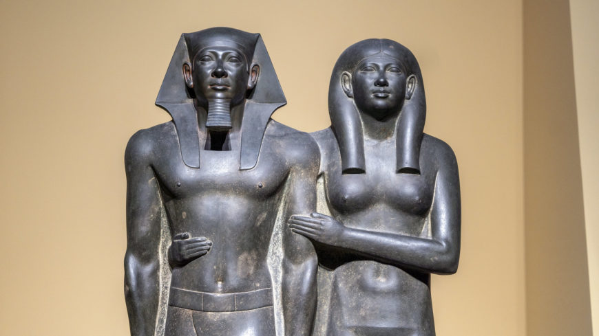 A photograph of Heads and torsos (detail), King Menkaure (Mycerinus) and queen statue standing side by side, depicting a male figure with a traditional headdress and a female figure with a close-fitting wig. Both statues have arms crossed over their chests and exhibit detailed craftsmanship with smooth, dark stone surfaces against a plain background.