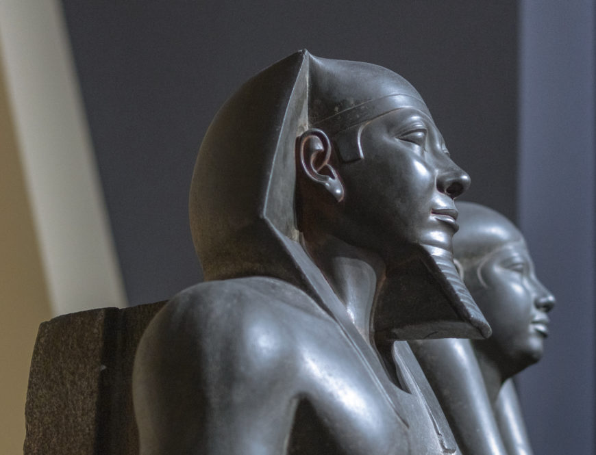 Photograph of Heads (detail), King Menkaure (Mycerinus) and queen stone statue depicting an ancient Egyptian figure wearing a traditional headdress. The statue features smooth, polished surfaces with detailed carving on the ears and headdress, set against a blurred background with soft lighting highlighting its contours.
