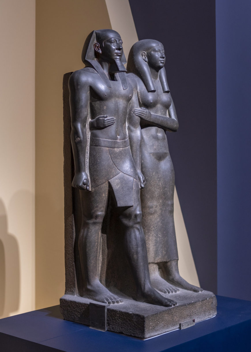 Photograph of King Menkaure (Mycerinus) and queen statue depicting a standing man and woman side by side, carved from dark stone with detailed features and traditional attire. The man wears a kilt and a headdress, while the woman wears a long dress, both standing on a rectangular base against a neutral background.