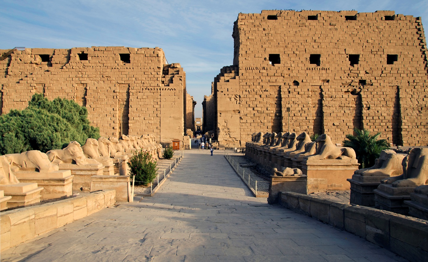 Photograph of sphinxes, the first pylon, and the central east-west aisle of Temple of Amon-Ra, Karnak in Luxor featuring large sandstone walls and rows of sphinx statues lining a wide stone pathway. The scene highlights architectural grandeur and historical significance with clear blue sky and sparse greenery framing the structure.