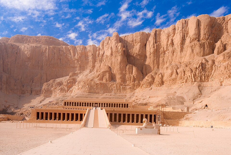Photograph of an ancient Egyptian temple complex set against a backdrop of rugged desert cliffs under a partly cloudy blue sky. The temple features a long central ramp leading to a colonnaded structure with multiple rectangular columns, flanked by a sphinx statue in the foreground.