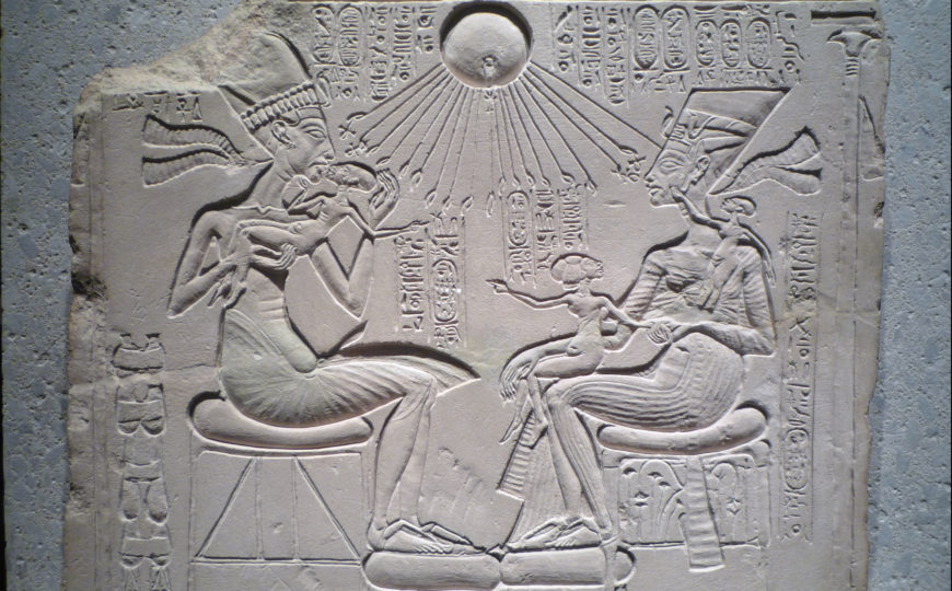 Bas-relief carving depicts two seated ancient Egyptian figures facing each other, with a child sitting on the lap of the figure on the right. Sun disk with rays ending in hands appears above, surrounded by hieroglyphic inscriptions.
