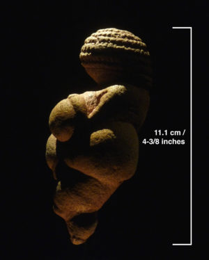 Photograph of a small ancient figurine measuring 11.1 cm (4-3/8 inches) tall, shown against a black background with a white measurement scale on the right. The figurine features a rounded head with textured hair and a stylized body with exaggerated curves.