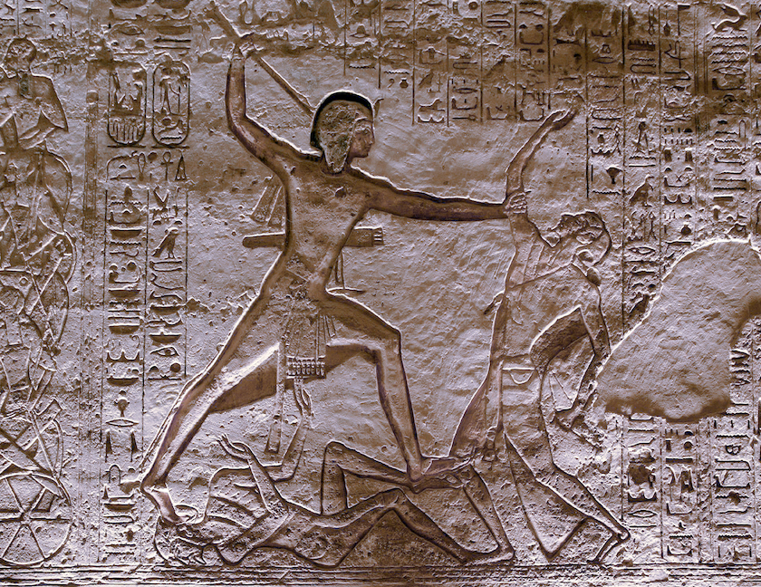 Bas-relief carving depicting an ancient Egyptian warrior in a dynamic pose, holding a weapon and subduing two enemies beneath him. Surrounding hieroglyphic inscriptions provide historical context, with detailed figures and symbolic elements carved into stone.