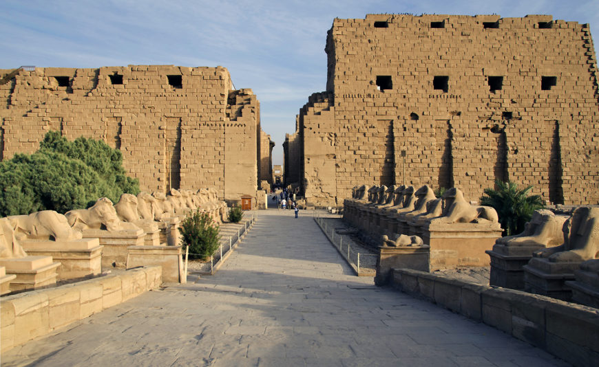 Photograph of sphinxes, the first pylon, and the central east-west aisle of Temple of Amon-Ra, Karnak in Luxor featuring large sandstone walls and rows of sphinx statues lining a wide stone pathway. The scene highlights architectural grandeur and historical significance with clear blue sky and sparse greenery framing the structure.