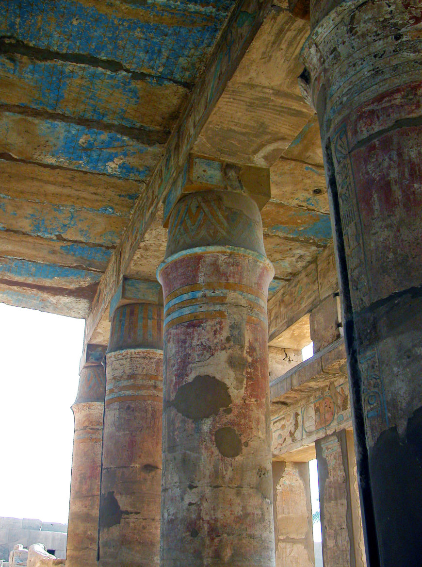 Photograph of ancient Egyptian columns inside a Karnak at Luxor showcasing faded red, blue, and beige paint with hieroglyphic carvings on the ceiling and walls. Columns feature distinct bands of color and weathered patches.