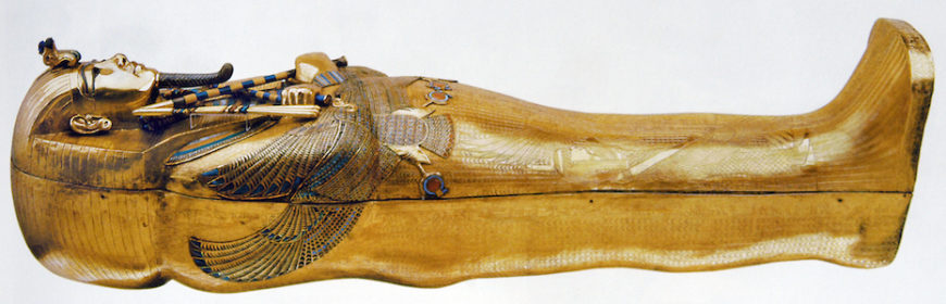 A photograph of Tutankhamun’s tomb, innermost coffin sarcophagus made of gold, featuring detailed engravings and colorful inlays on the chest and head area. The sarcophagus showcases traditional Egyptian artistic elements, including a headdress, crossed arms, and intricate wing designs symbolizing protection.