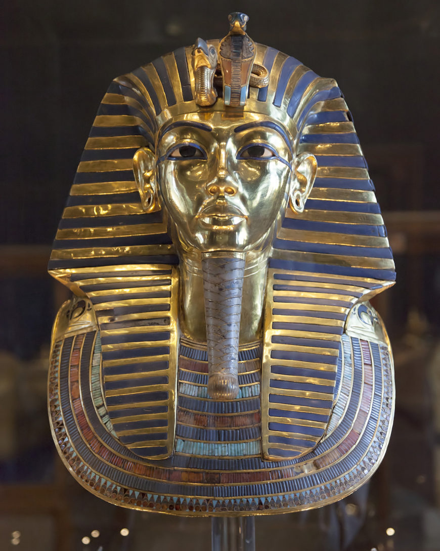 Photograph of Death Mask from innermost coffin, Tutankhamun’s tomb, featuring gold and blue striped headdress with intricate inlays of colored stones along the collar. The mask displays a stylized beard and cobra emblem on the forehead, symbolizing royalty and divine protection.