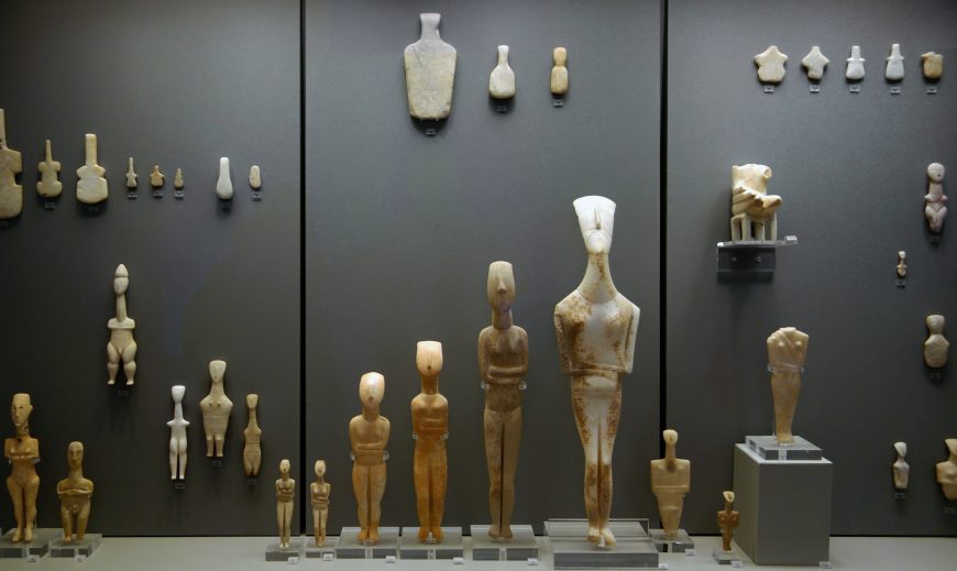 Photograph of a museum display featuring various ancient Cycladic figurines arranged by size and shape against a dark background. Figurines range from small, detailed busts to larger, full-body sculptures.