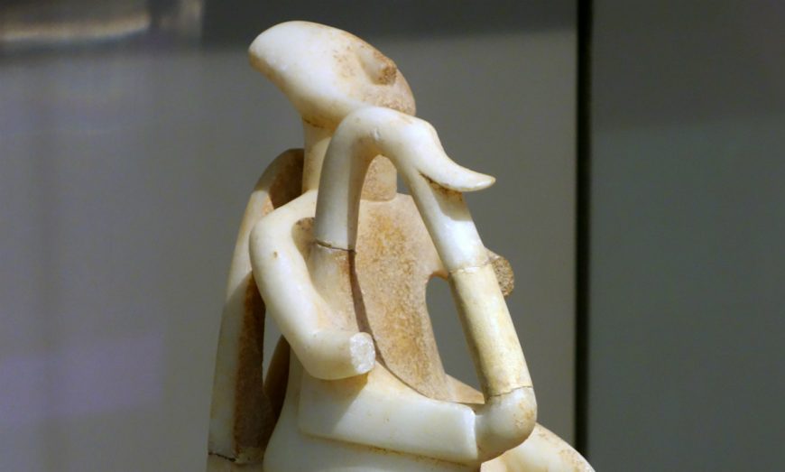 Photograph of an ancient marble Cycladic sculpture depicting a seated figure playing a harp, showcasing detailed carving and smooth texture. The sculpture highlights curved arms, a rounded head, and a harp with visible strings.