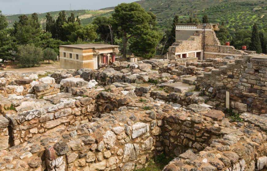 Photograph of ancient archaeological site at Knossos with stone walls and partially reconstructed buildings set against a backdrop of green hills and trees. The site features notable restored structures with red and white painted details, highlighting historical architecture and excavation efforts.