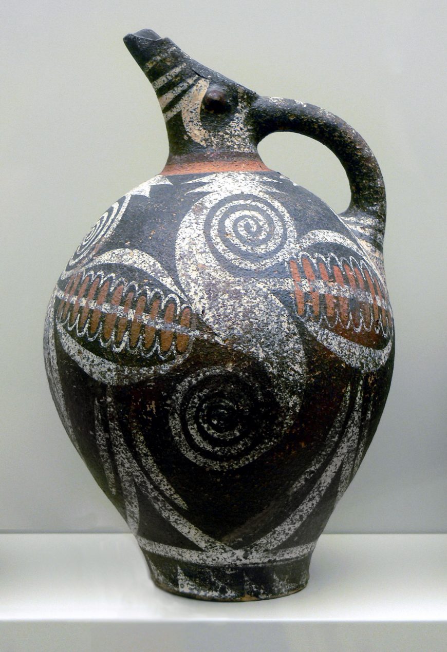 Photograph of Kamares ware jug from Phaistos with a rounded body, narrow neck, and handle, decorated with intricate spiral and geometric patterns in black, white, and reddish-brown. Jug features a spout shaped like an animal head, showcasing artistic craftsmanship and cultural symbolism.