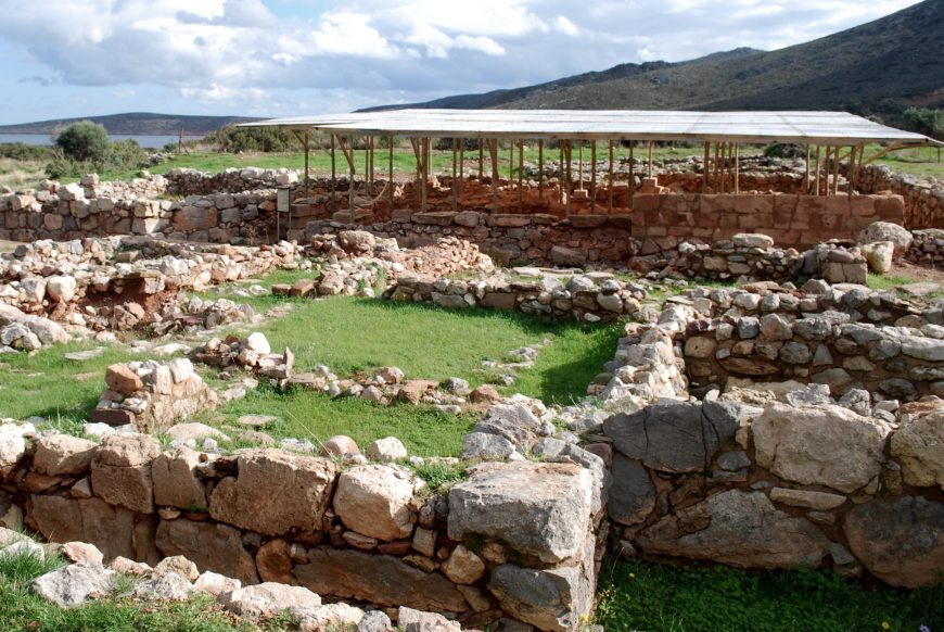Photograph of ancient stone ruins at Palaikastro with multiple low walls forming rectangular enclosures, partially covered by a large metal roof supported by wooden poles. The site is set in a grassy area with hills in the background, indicating an archaeological excavation of a historic settlement.