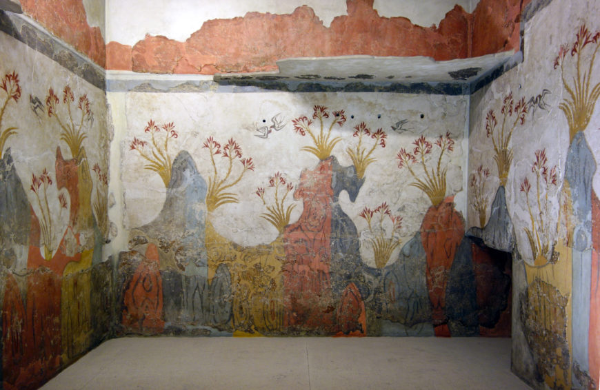 Photograph of an ancient fresco mural covering three walls of a small room, depicting stylized plants and figures in red, yellow, and gray tones. The artwork features tall floral motifs with elongated stems and clusters of flowers, set against a white background with a red border near the ceiling, illustrating decorative and symbolic elements from historical interior design.