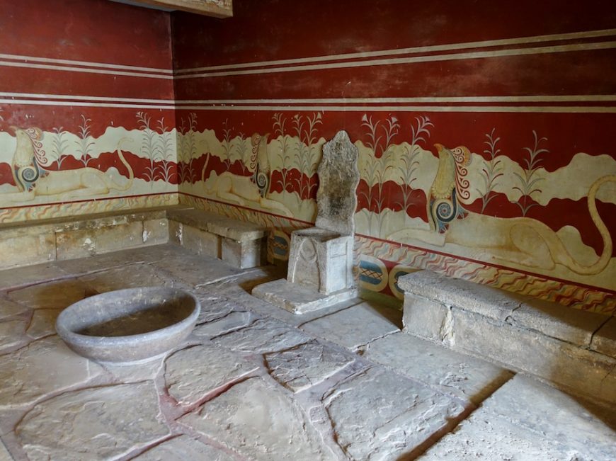 Photograph of Throne Room, Knossos with stone benches, a carved stone chair, and a large circular basin on a stone floor. Walls feature red background with decorative white and yellow frescoes of lions and floral patterns.