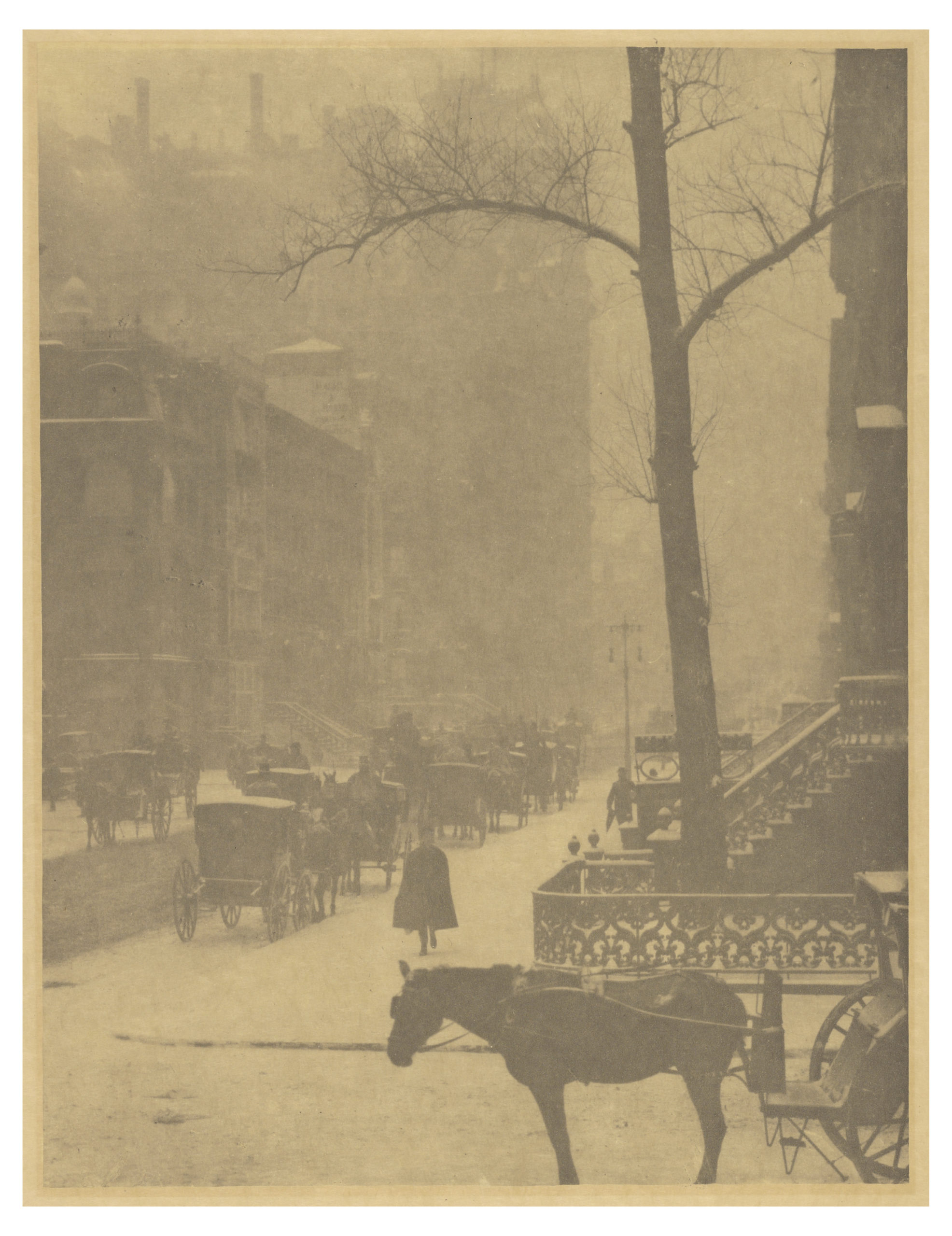 Photograph of a foggy urban street scene from the early 20th century, featuring horse-drawn carriages, a pedestrian, and leafless trees along a sidewalk. The sepia-toned view highlights historic architecture, street activity, and atmospheric conditions, emphasizing transportation and daily life of the era.