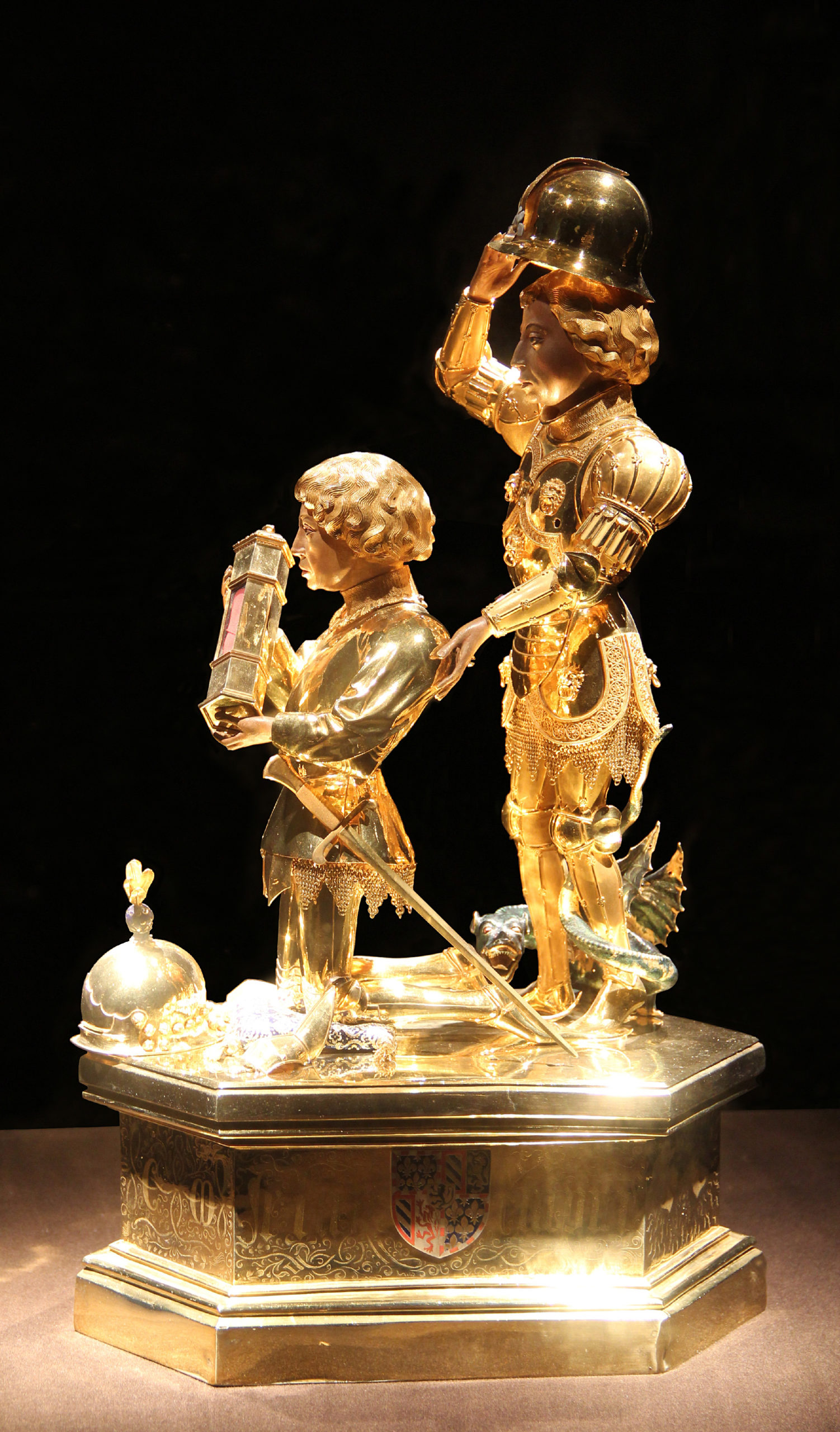 Photograph of an ornate golden sculpture featuring two figures, one holding a lantern and the other lifting a helmet, both standing on a detailed hexagonal base with an engraved crest. The sculpture showcases intricate armor and decorative elements.