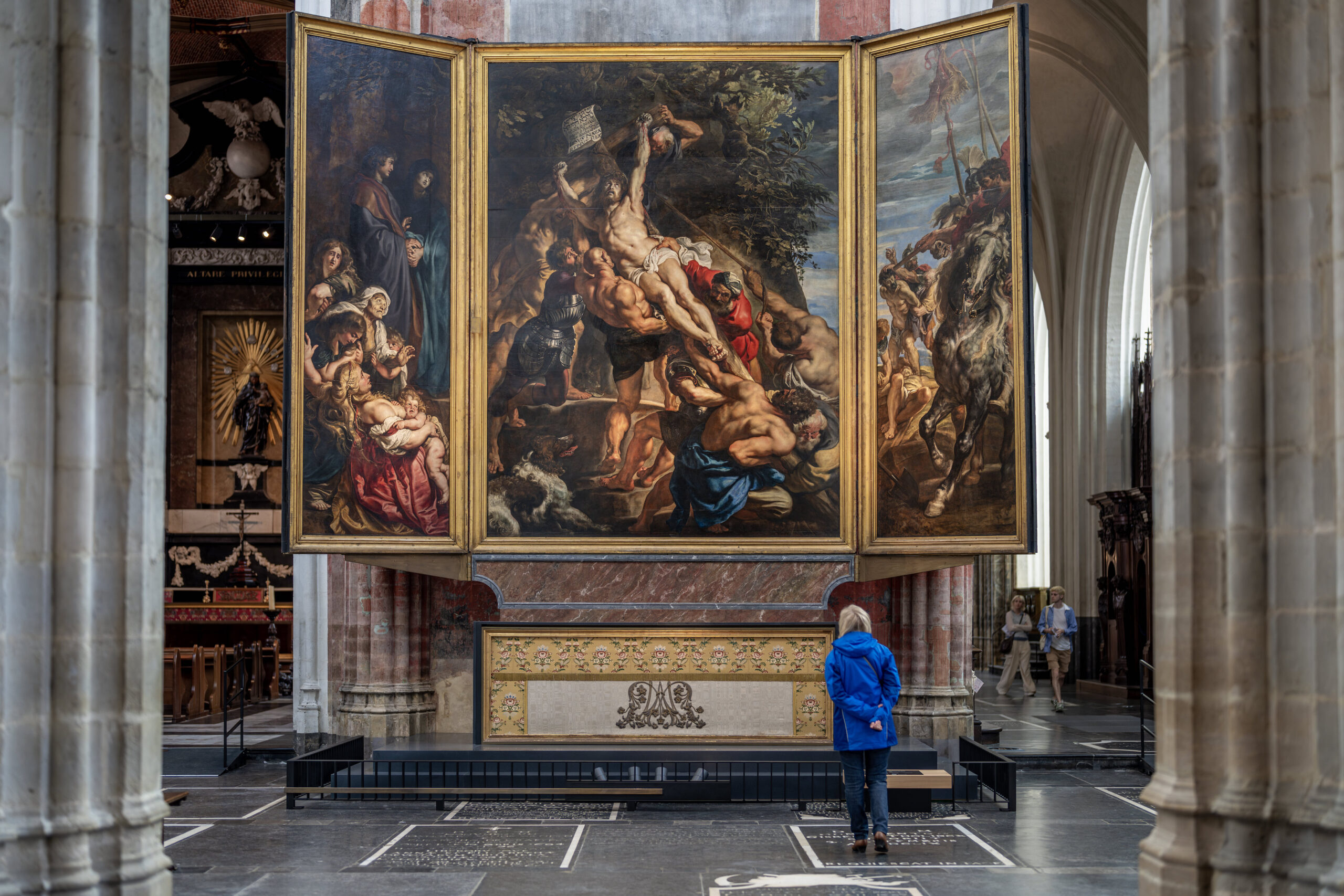 Photograph of a large triptych painting displayed inside a cathedral, depicting religious scenes with multiple human figures in dramatic poses and rich colors. A person in a blue jacket stands in front, highlighting scale, while architectural columns and arched windows frame the artwork.