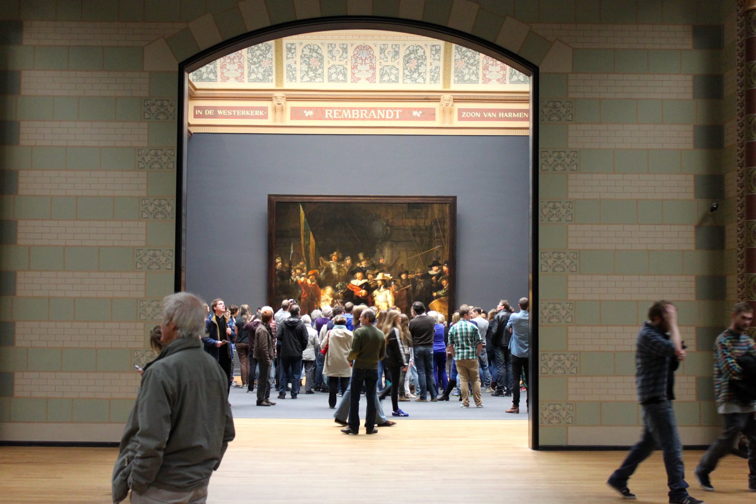 Photograph of a museum gallery showing a group of people gathered around a large framed painting by Rembrandt. The scene highlights the painting's central position on a dark wall with an arched doorway framing the view and visitors observing or walking nearby.
