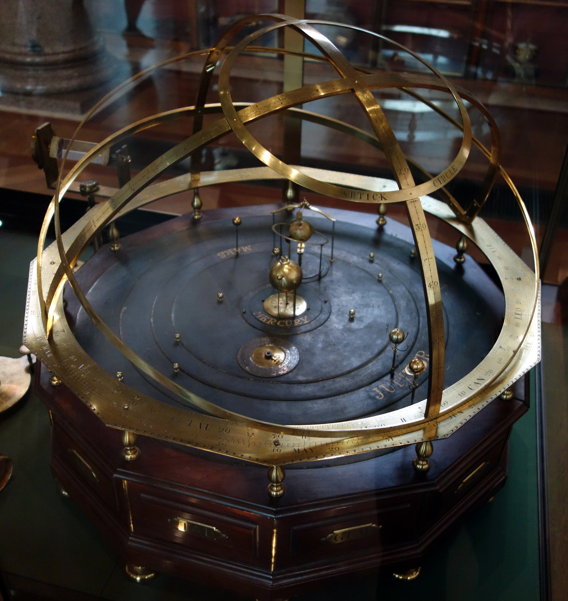 Photograph of an antique orrery model demonstrating heliocentric solar system with brass rings and spheres representing planets orbiting a central sun. Features engraved circular bands with celestial markings and a wooden base with drawers, highlighting historical astronomical study tools.