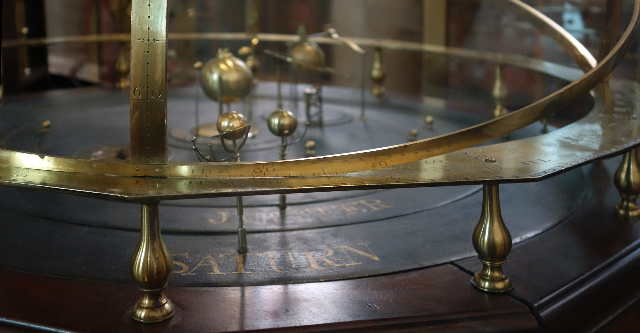 Photograph of an antique orrery model demonstrating heliocentric solar system with brass rings and spheres representing planets orbiting a central sun. Features engraved circular bands with celestial markings and a wooden base with drawers, highlighting historical astronomical study tools.