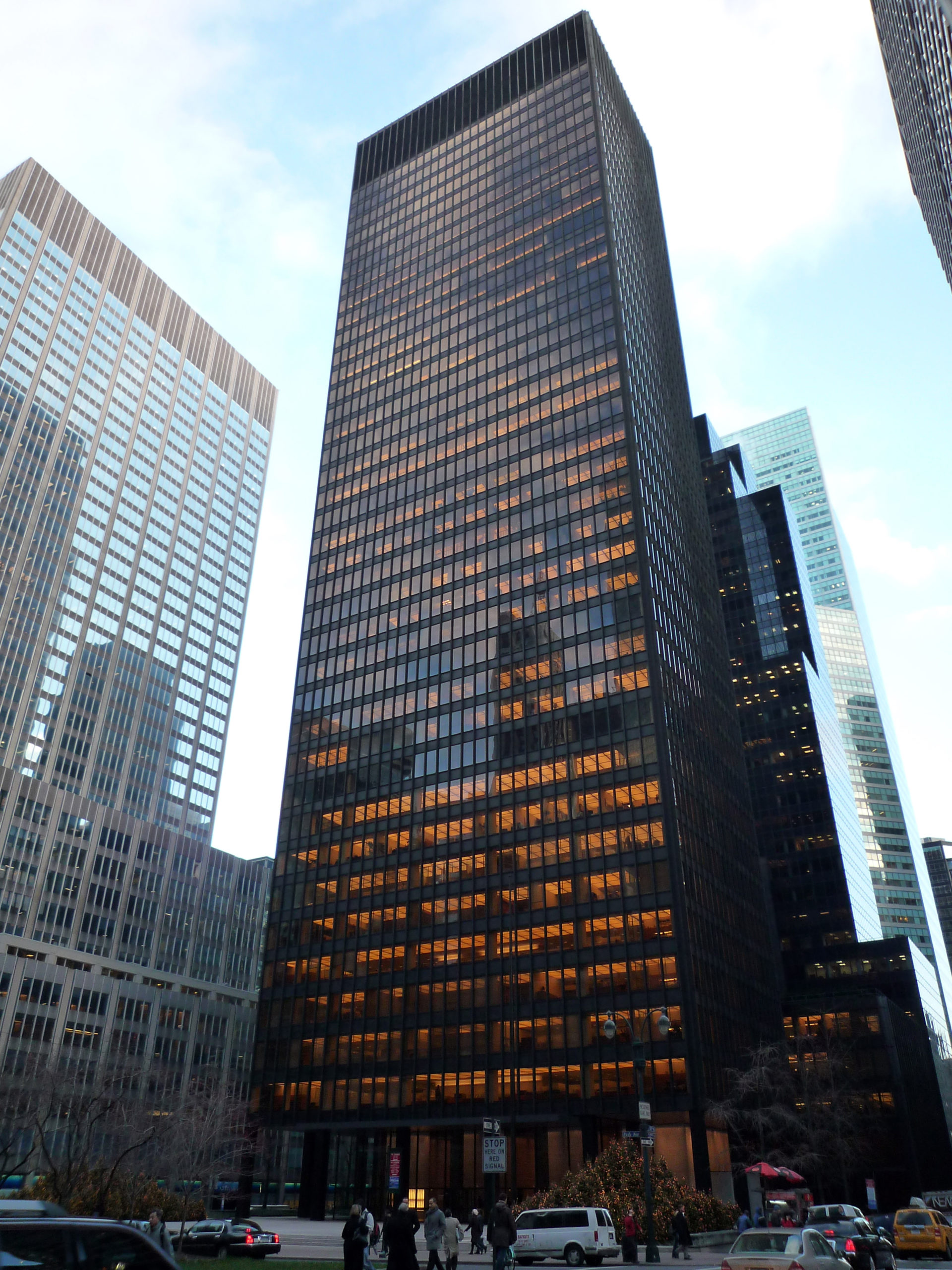 Photograph of a tall, dark glass skyscraper reflecting orange sunlight, surrounded by other high-rise buildings in a cityscape. The building's grid-like window pattern and street-level activity with cars and pedestrians highlight an urban business district during late afternoon.