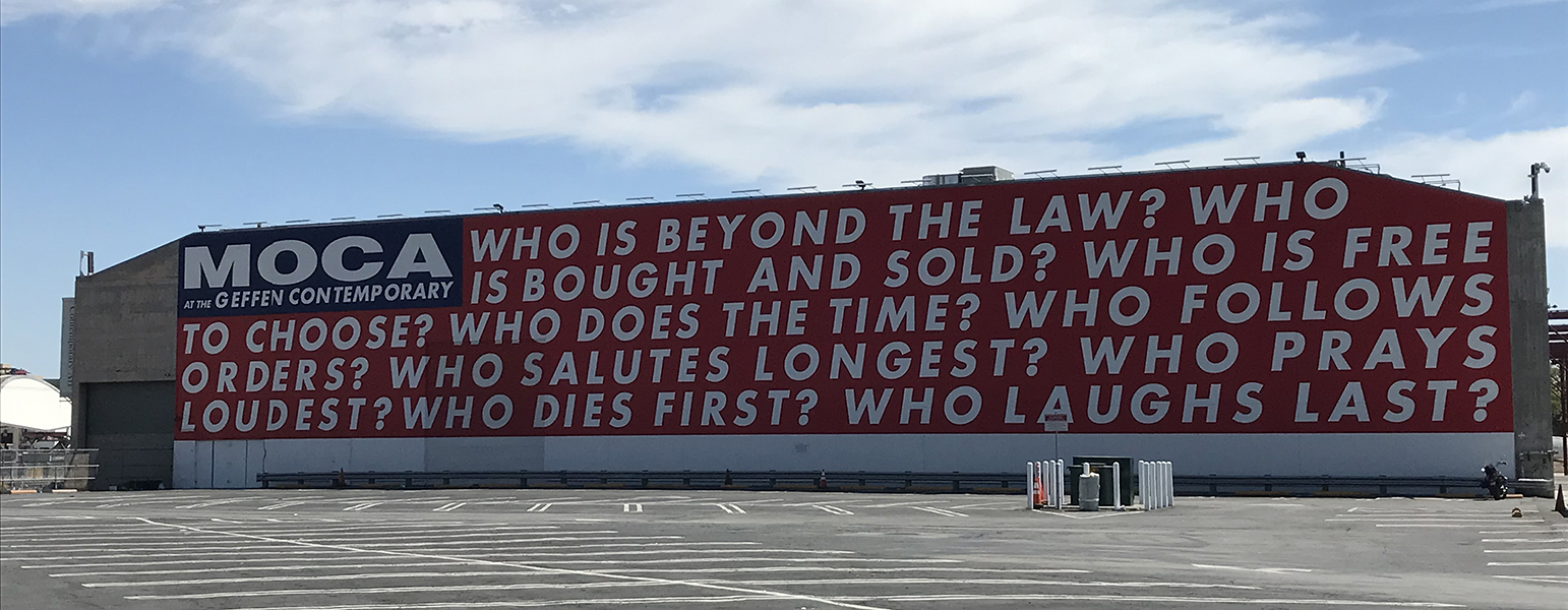 Photograph of a large mural on a building wall featuring bold white text on a red background with "MOCA" and "Geffen Contemporary" in black and white on the left side. The mural poses a series of provocative questions about law, freedom, loyalty, and mortality.