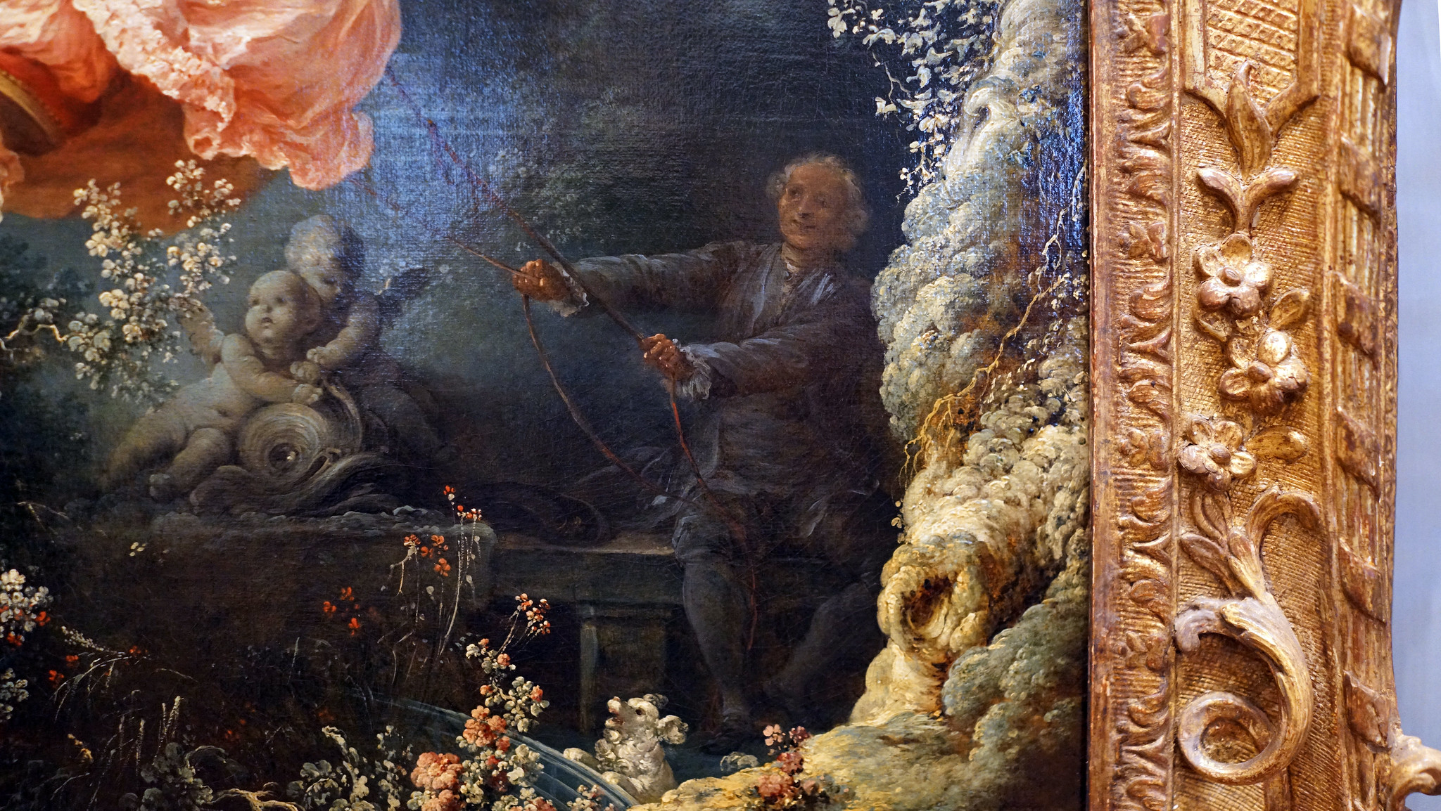 A photograph of a detail of Fragonard's The Swing, with a figure holding the strings to a swing surrounded by intricate floral and cherubic details. The painting is framed with an ornate gold border decorated with floral and scroll motifs.