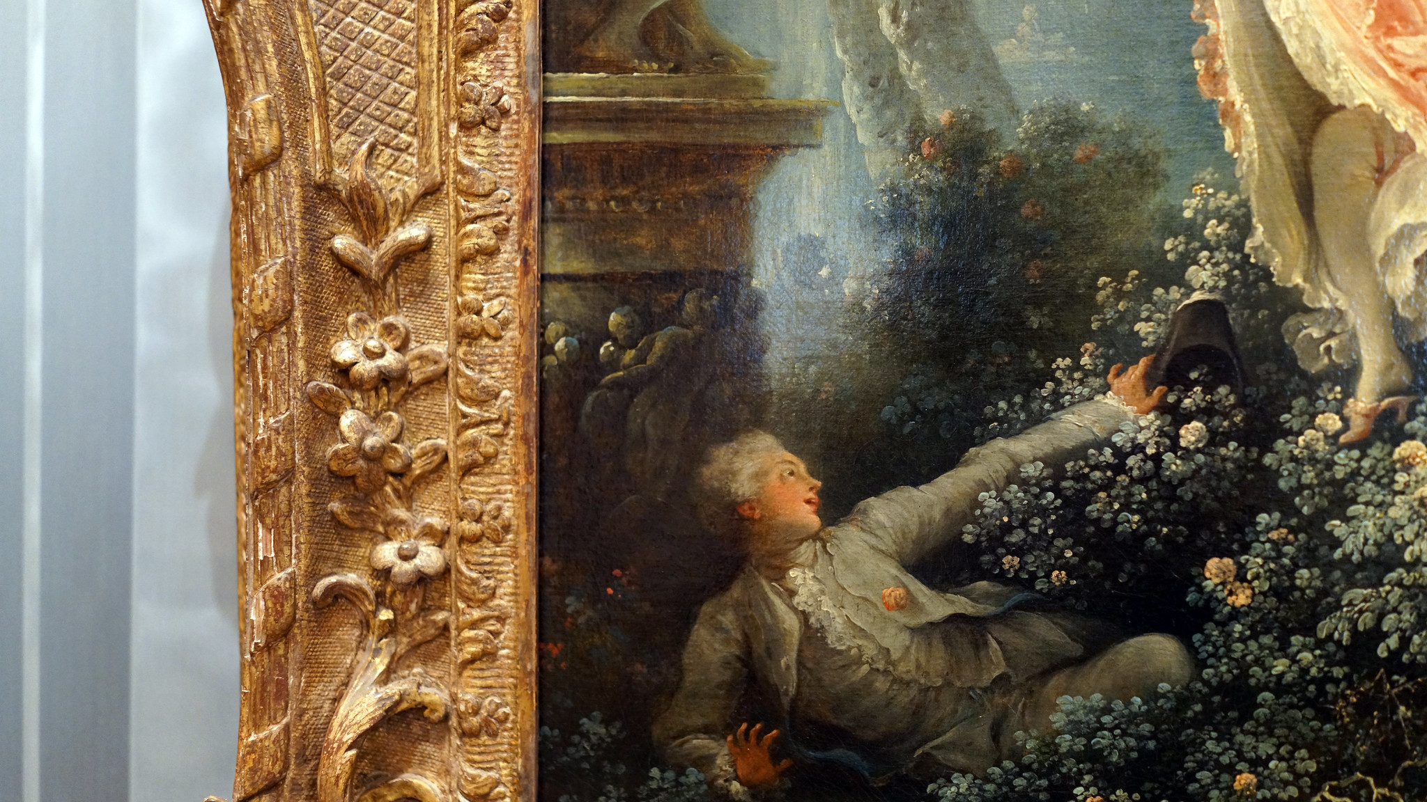 Photograph of a detailed section of Fragonard's The Swing featuring a reclining figure in 18th-century attire surrounded by dense foliage and flowers. Ornate golden frame with floral and geometric patterns is visible on the left side.