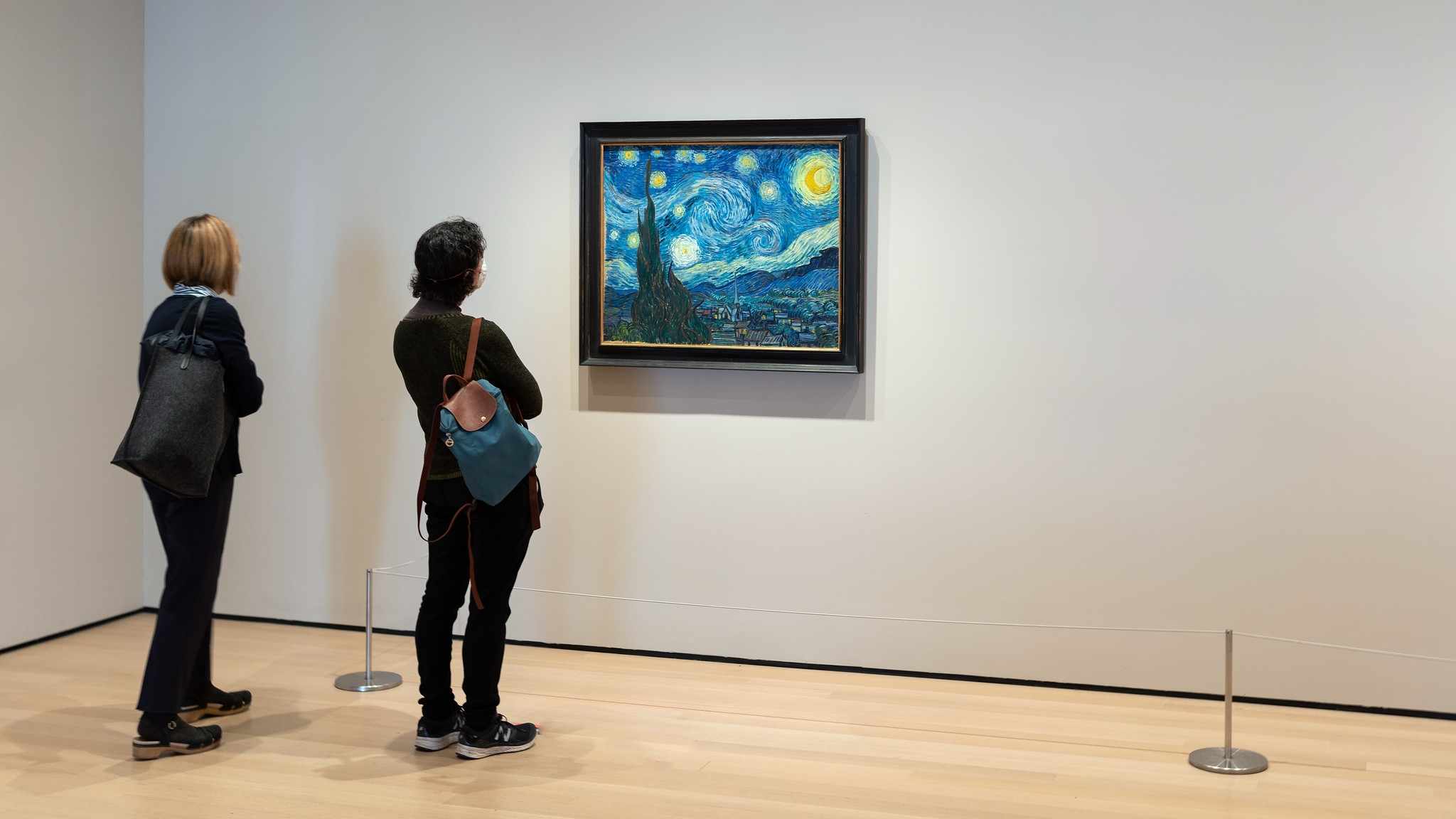 Photograph showing two people standing in an art gallery observing Vincent van Gogh's "Starry Night" painting. The painting features swirling blue and yellow night sky over a quiet village, framed and mounted on a white wall with wooden flooring and minimalistic barriers around.