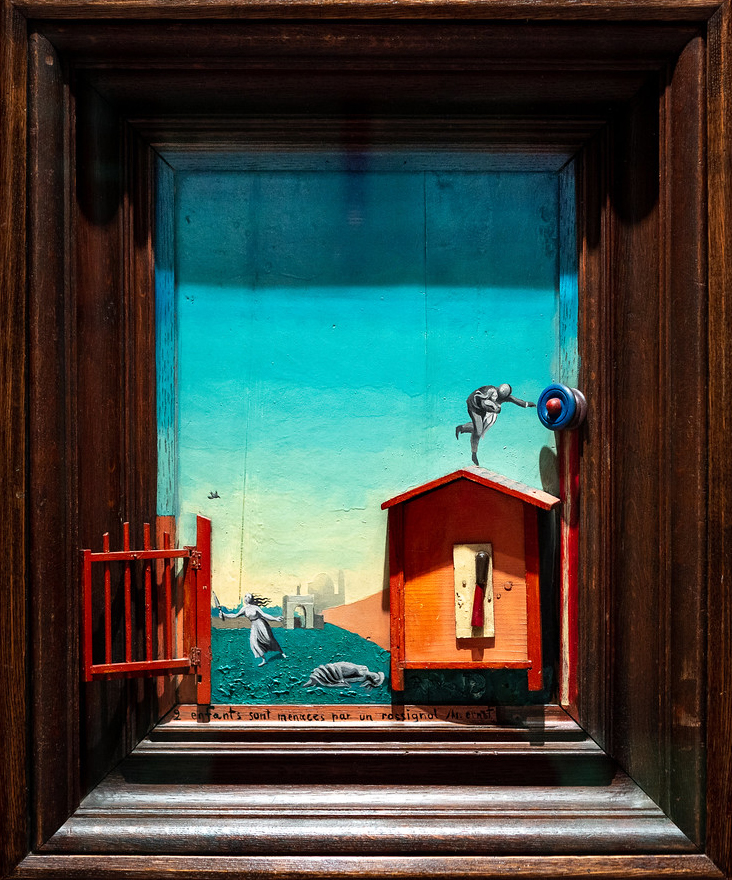 Photograph of a framed miniature diorama depicting a scene with a bright blue sky. The scene includes a small orange shed, a red gate, a bird perched on a pole, and several figures on the ground.