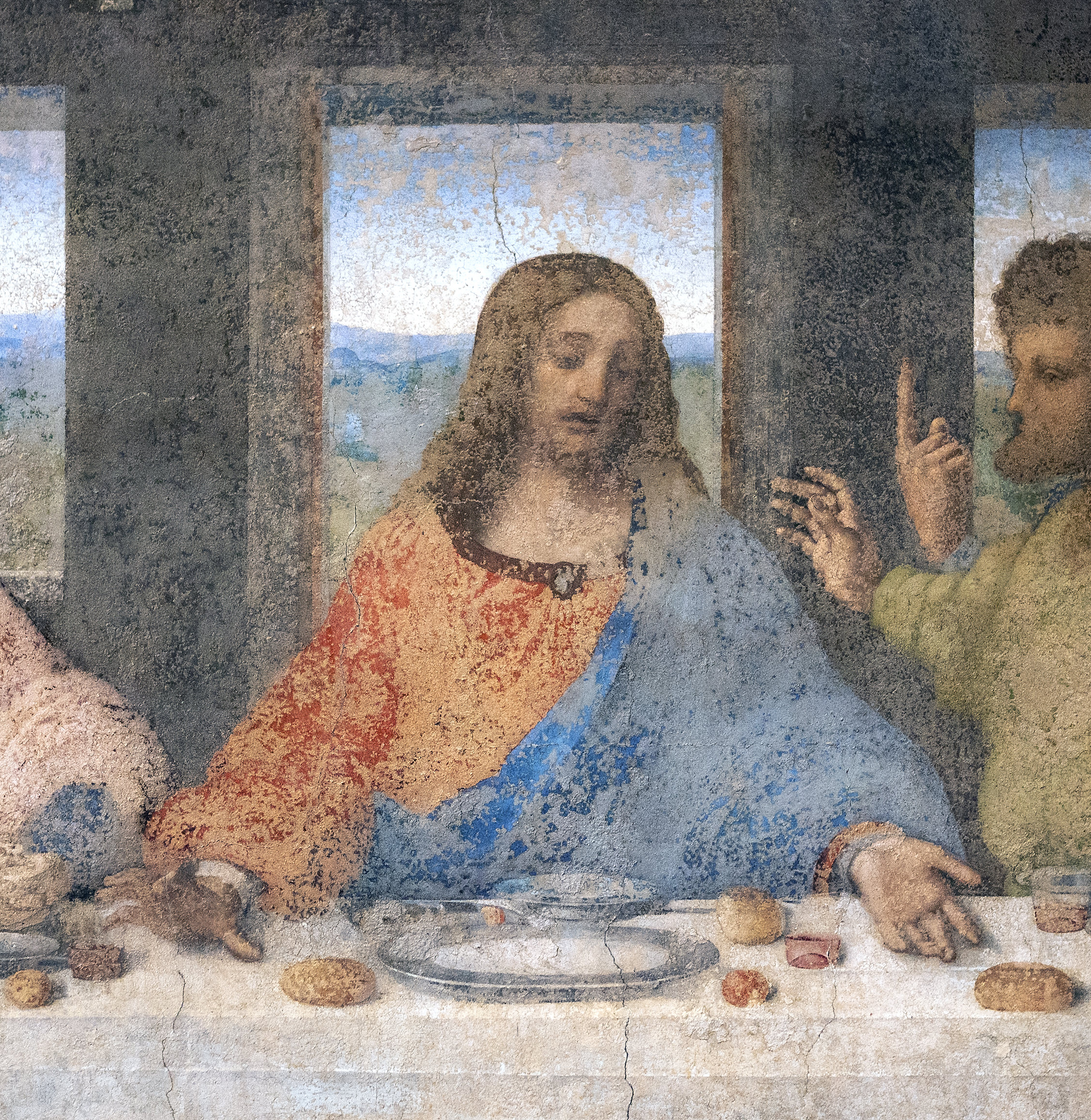 Christ (detail), Leonardo da Vinci, Last Supper. The central figure wears a red and blue robe, with bread and cups placed on the table, set against a backdrop of windows showing a distant landscape.