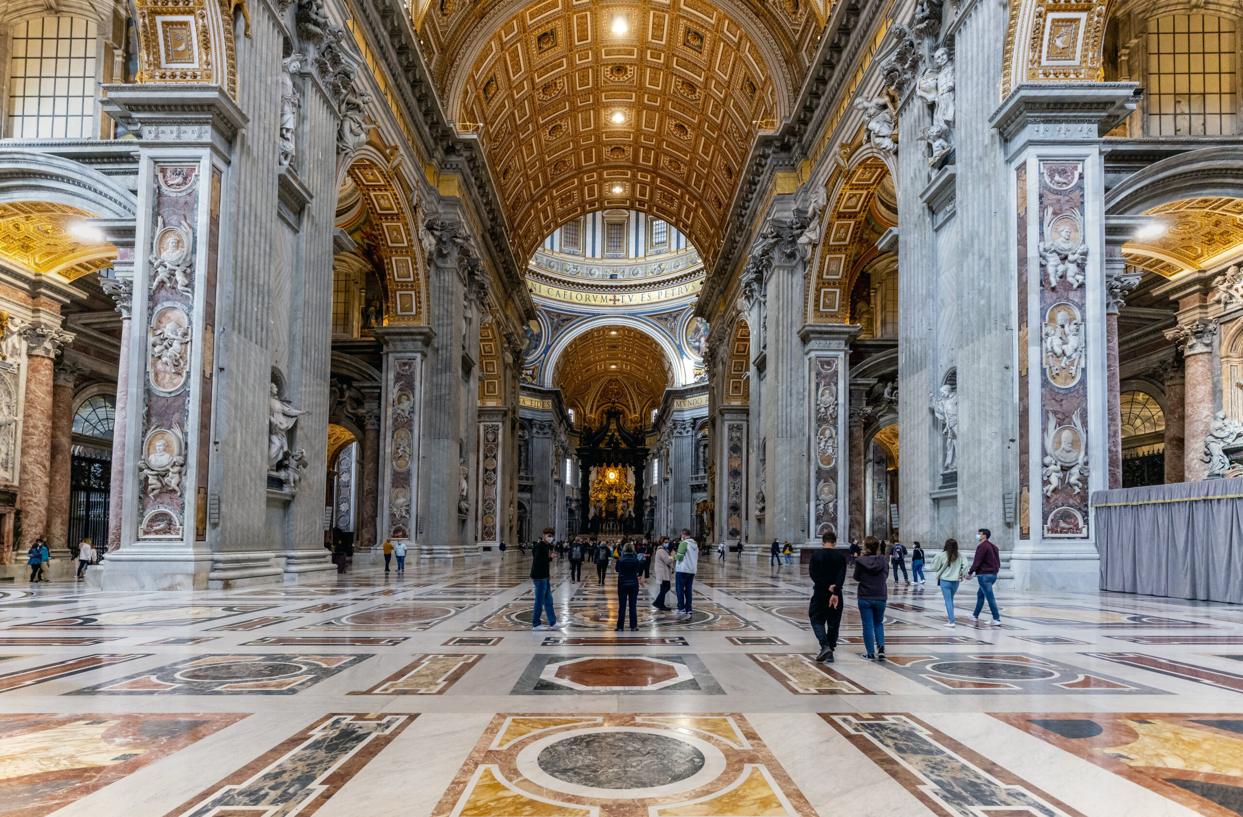Photograph interior of Saint Peter's Basilica, Vatican City featuring a high vaulted ceiling with intricate gold detailing and large marble columns adorned with sculptures and artwork. Visitors walk across a polished, patterned marble floor, highlighting the vast scale and ornate architectural design.