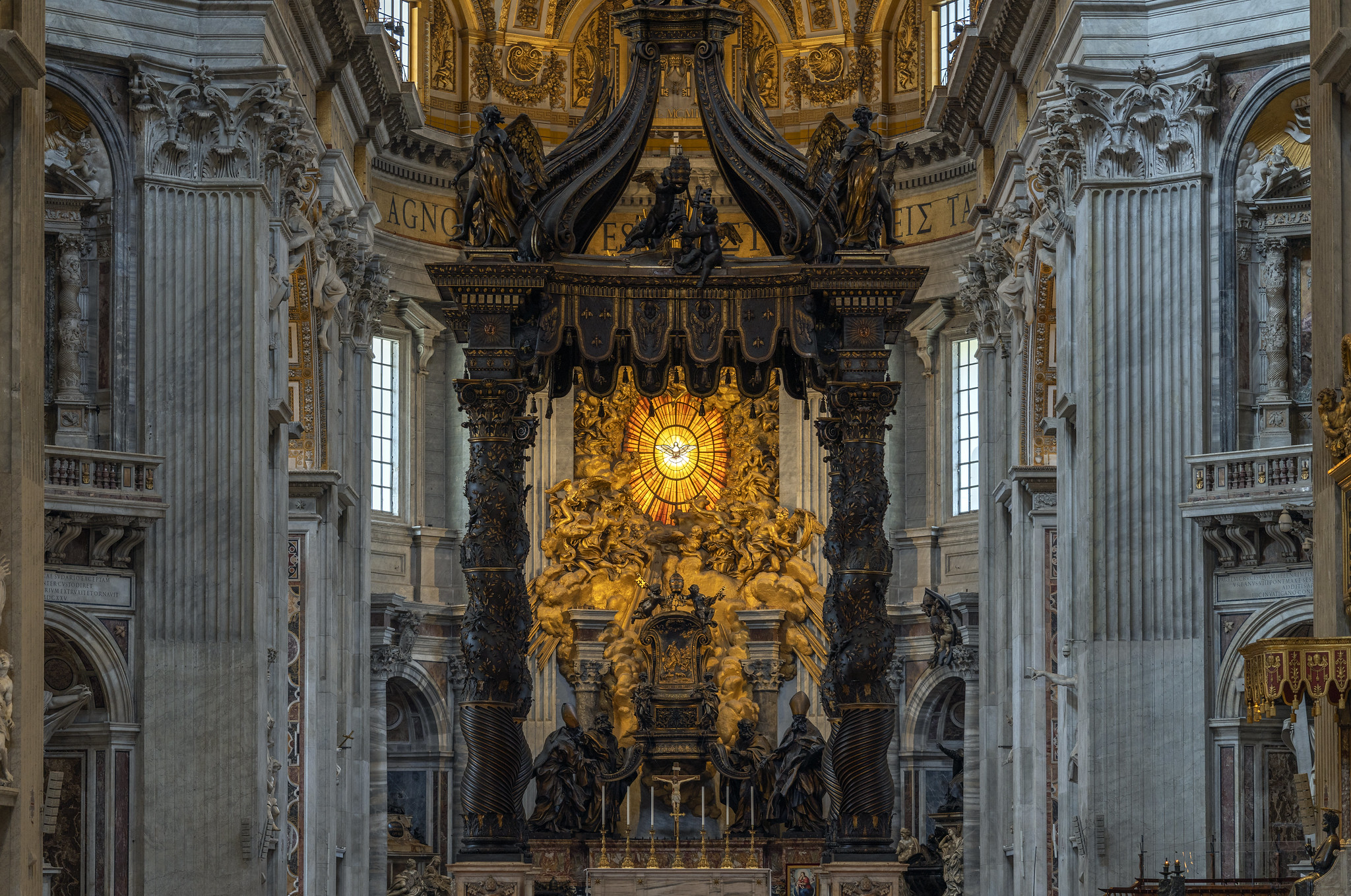 Photograph of Gian Lorenzo Bernini, View to Cathedra Petri (or Chair of St. Peter) showing an ornate altar inside a grand cathedral, featuring a central sculptural group illuminated by a glowing circular stained glass window with a dove symbol. The altar is framed by four large twisted columns and surrounded by intricate architectural details, including tall marble pillars and elaborate gold decorations on the ceiling