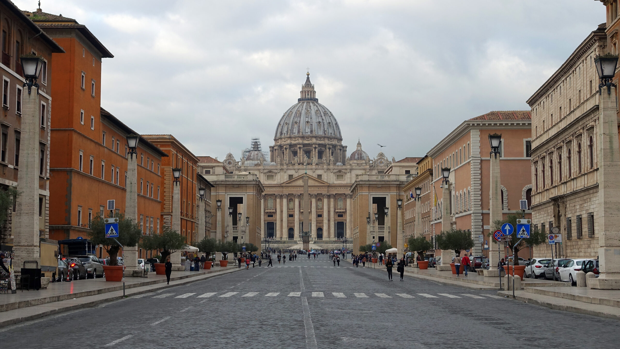 Photograph of a wide street leading to St. Peter's Basilica in Vatican City, showcasing Renaissance architecture and a large dome under a cloudy sky. The street is lined with historic buildings, street lamps, and pedestrians, highlighting a calm urban scene.