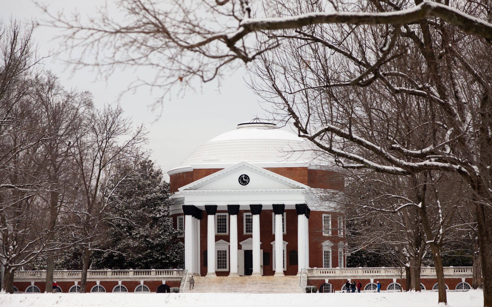Photograph of Thomas Jefferson rotunda building with a white dome and tall white columns, surrounded by leafless trees dusted with snow. The scene captures a winter setting with a few people visible near the entrance.