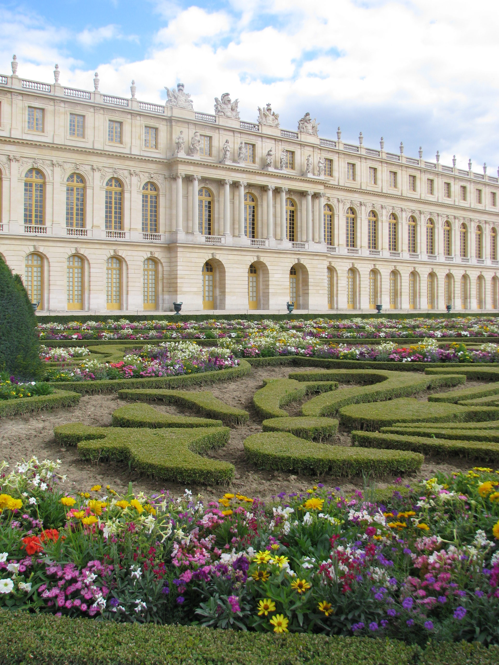 Photograph of Versailles with symmetrical architecture featuring tall arched windows and ornate sculptures along the roofline. In the foreground, a formal garden displays neatly trimmed hedges arranged in intricate patterns surrounded by colorful blooming flowers under a partly cloudy sky.